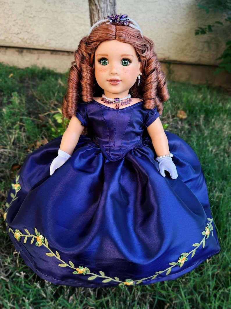 Puede incluir: Mu&ntilde;eca con cabello casta&ntilde;o rojizo y ojos verdes, vestida con un vestido de sat&eacute;n azul oscuro con bordados florales. La mu&ntilde;eca lleva una tiara y un collar, y est&aacute; sobre hierba verde.