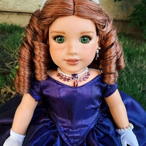 Puede incluir: Mu&ntilde;eca con cabello casta&ntilde;o rojizo, ojos verdes y pecas, vestida con un vestido de sat&eacute;n azul oscuro, guantes blancos y joyas de perlas. Una tiara enjoyada adorna el cabello de la mu&ntilde;eca. La mu&ntilde;eca posa al aire libre sobre una superficie cubierta de hierba.