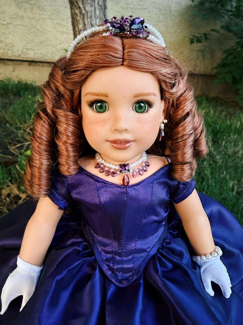 Puede incluir: Mu&ntilde;eca con cabello casta&ntilde;o rojizo, ojos verdes y pecas, vestida con un vestido de sat&eacute;n azul oscuro, guantes blancos y joyas de perlas. Una tiara enjoyada adorna el cabello de la mu&ntilde;eca. La mu&ntilde;eca posa al aire libre sobre una superficie cubierta de hierba.