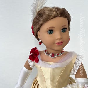 May include: A doll in a cream-colored dress with a red and pearl necklace and earrings. The doll has a white feather in its hair and is holding a fan. The dress has red flower accents and white lace trim.