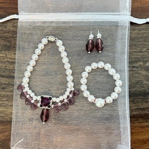 Puede incluir: Un conjunto de joyas que incluye un collar, una pulsera y pendientes, presentado en una bolsa de organza blanca. El collar y la pulsera est&aacute;n hechos de perlas blancas de imitaci&oacute;n, con cuentas moradas y un colgante cuadrado morado. Los pendientes tienen forma de l&aacute;grima.