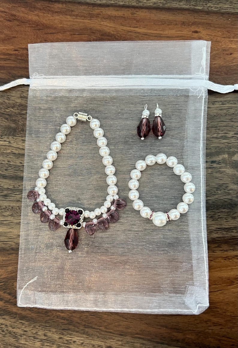 Puede incluir: Un conjunto de joyas que incluye un collar, una pulsera y pendientes, presentado en una bolsa de organza blanca. El collar y la pulsera est&aacute;n hechos de perlas blancas de imitaci&oacute;n, con cuentas moradas y un colgante cuadrado morado. Los pendientes tienen forma de l&aacute;grima.