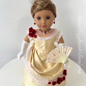 Puede incluir: Mu&ntilde;eca con un vestido color crema con detalles florales rojos, un collar y pendientes a juego. La mu&ntilde;eca tiene un adorno para el pelo con plumas, guantes blancos y un abanico decorativo. El vestido de la mu&ntilde;eca recuerda a la moda hist&oacute;rica.