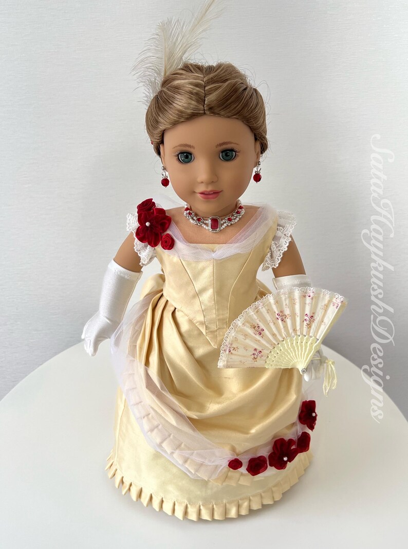 Puede incluir: Mu&ntilde;eca con un vestido color crema con detalles florales rojos, un collar y pendientes a juego. La mu&ntilde;eca tiene un adorno para el pelo con plumas, guantes blancos y un abanico decorativo. El vestido de la mu&ntilde;eca recuerda a la moda hist&oacute;rica.