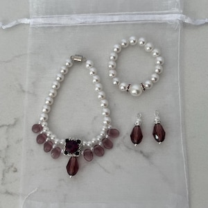 Puede incluir: Un conjunto de joyas con un collar de perlas, una pulsera y pendientes. El collar tiene una pieza central cuadrada de color rojo oscuro y detalles en forma de l&aacute;grima. La pulsera y los pendientes tienen detalles en rojo oscuro a juego. El conjunto est&aacute; en una bolsa blanca transparente.