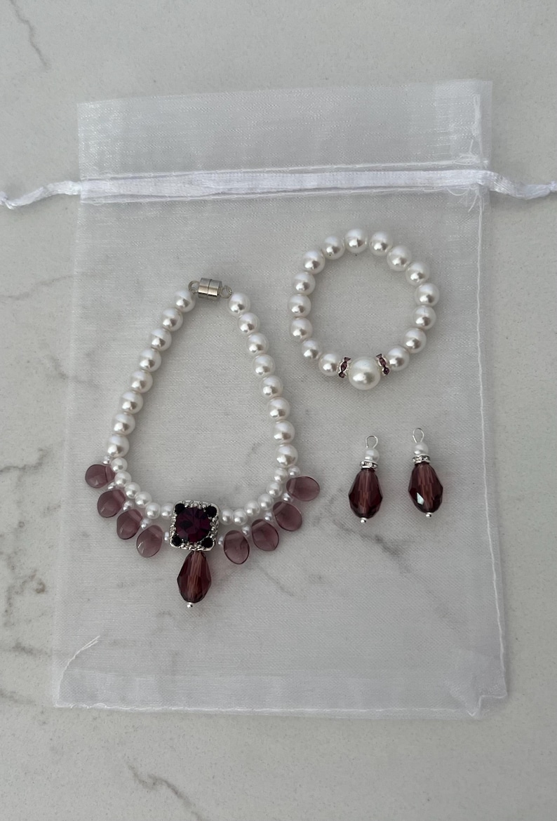 Puede incluir: Un conjunto de joyas con un collar de perlas, una pulsera y pendientes. El collar tiene una pieza central cuadrada de color rojo oscuro y detalles en forma de l&aacute;grima. La pulsera y los pendientes tienen detalles en rojo oscuro a juego. El conjunto est&aacute; en una bolsa blanca transparente.