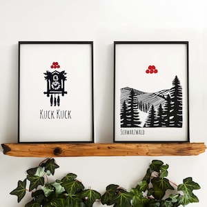 May include: Two black framed prints on a wooden shelf. The print on the left features a black and white illustration of a cuckoo clock with the text "KUCK KUCK". The print on the right features a black and white illustration of a mountain range with the text "SCHWARZWALD".