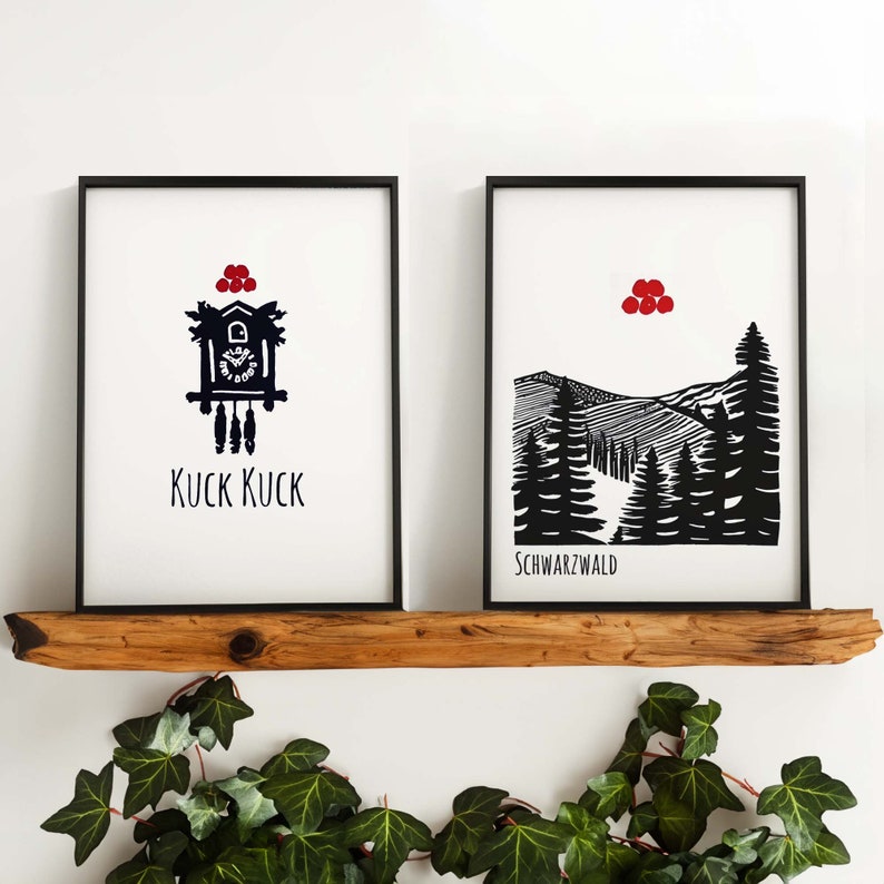 May include: Two black framed prints on a wooden shelf. The print on the left features a black and white illustration of a cuckoo clock with the text "KUCK KUCK". The print on the right features a black and white illustration of a mountain range with the text "SCHWARZWALD".