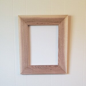 May include: A simple, rectangular wooden picture frame with a light brown finish. The frame is hanging on a white wall.