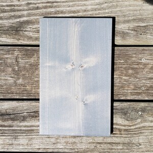 May include: A rectangular wooden sign, painted in a pale blue hue. The wood grain is visible, and the sign has a smooth surface. It is displayed on a weathered wooden surface, showing natural wood grain.