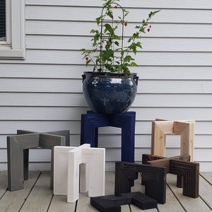 Op de afbeelding: Houten plantenstandaards in diverse kleuren: grijs, wit, marineblauw, natuurlijk hout, zwart en bruin. Een blauwe keramische pot met plant en trellis staat op &eacute;&eacute;n standaard. De standaards hebben een geometrisch, modern ontwerp.