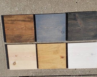 May include: A collection of six square wooden samples, each with a different stain. The top row features dark brown, gray, and black stains. The bottom row displays light wood, golden, and white finishes. The samples are arranged on a concrete surface.