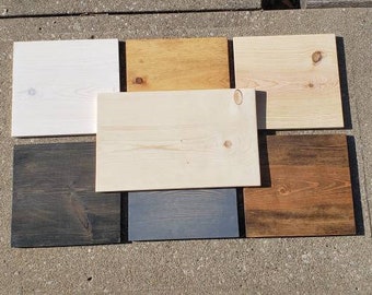 May include: Seven wood samples in various shades of brown, black, gray, and white. The samples are arranged in two rows on a concrete surface.