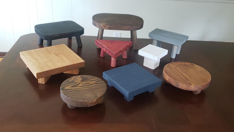 May include: A collection of nine wooden stools in various colors and styles. The stools are made of different types of wood and have a variety of finishes. Some of the stools are square, while others are round. The stools are all different sizes and heights.