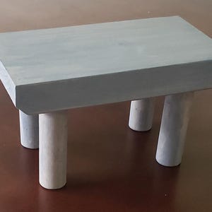 May include: A small, gray wooden stool with four legs. The stool is made of rough-hewn wood and has a rustic, farmhouse style.