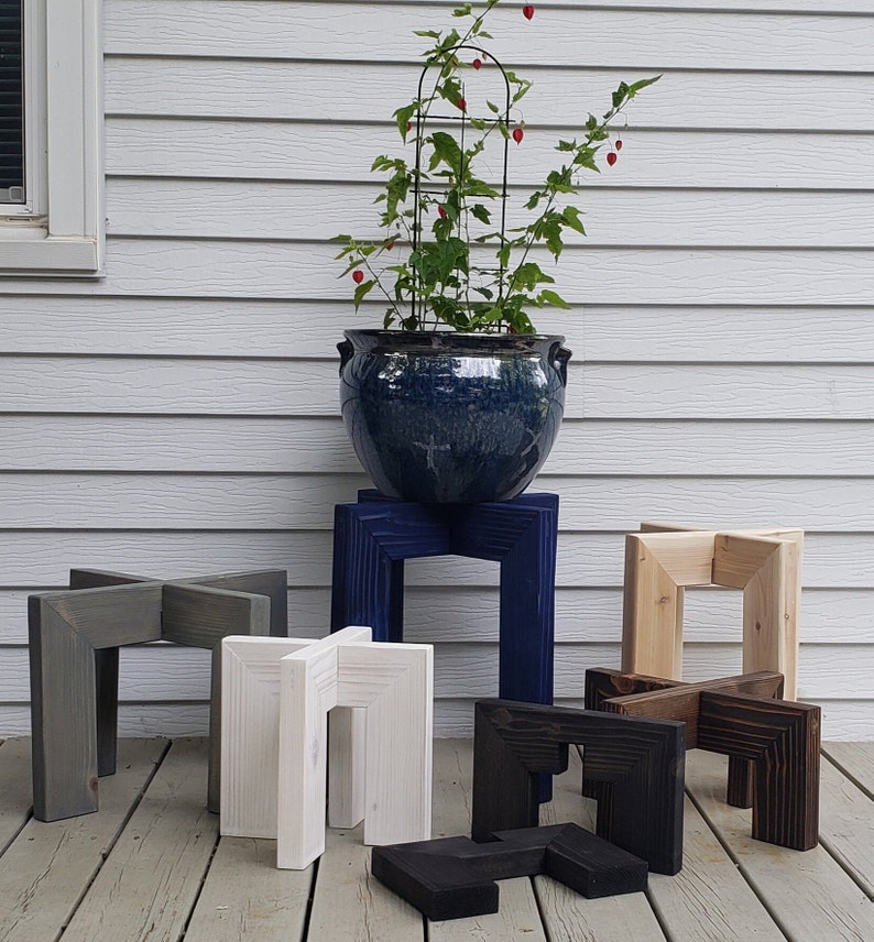 Op de afbeelding: Houten plantenstandaards in diverse kleuren: grijs, wit, marineblauw, natuurlijk hout, zwart en bruin. Een blauwe keramische pot met plant en trellis staat op &eacute;&eacute;n standaard. De standaards hebben een geometrisch, modern ontwerp.