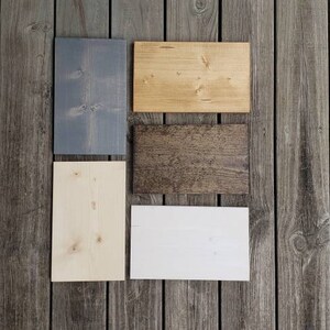 May include: Five rectangular wooden planks in shades of grey, light brown, dark brown, white, and natural wood. The planks are arranged on a weathered grey wooden surface, demonstrating different wood finishes and textures.