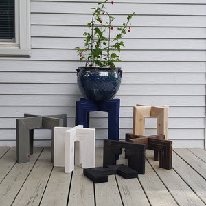 Op de afbeelding: Een verzameling houten plantenstandaards in verschillende kleuren, waaronder grijs, wit, blauw, natuurlijk hout, zwart en donkerbruin. De standaards worden getoond op een houten terras, met een grote blauwe keramische pot met een plant op de achtergrond.