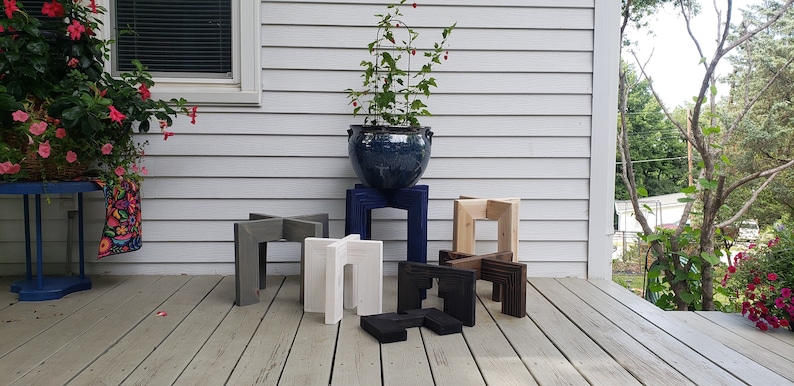 Op de afbeelding: Een verzameling houten plantenstandaards in verschillende kleuren, waaronder grijs, wit, blauw, natuurlijk hout, zwart en donkerbruin. De standaards worden getoond op een houten terras, met een grote blauwe keramische pot met een plant op de achtergrond.