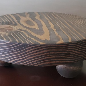 May include: A round wooden plant stand with a dark brown wood grain finish. The stand has three legs and a slightly raised center.