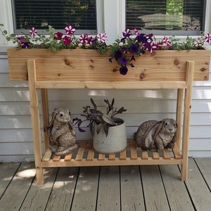 May include: A wooden planter box with a shelf underneath. The planter box is filled with pink and purple flowers. Two small rabbit statues are on the shelf.