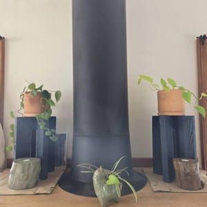 Op de afbeelding: Een interieursc&egrave;ne met een donkergrijze kachelpijp en twee donkerblauwe houten plantenstandaards. Elke standaard bevat een terracotta pot met hangende groene planten. Decoratieve houten en stenen accenten bevinden zich aan de basis.