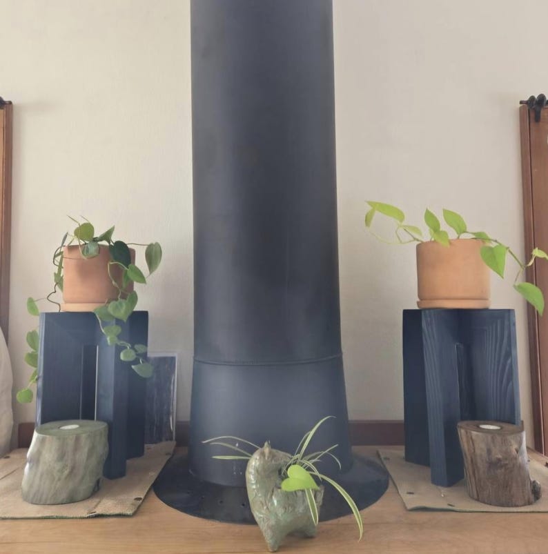 Op de afbeelding: Een interieursc&egrave;ne met een donkergrijze kachelpijp en twee donkerblauwe houten plantenstandaards. Elke standaard bevat een terracotta pot met hangende groene planten. Decoratieve houten en stenen accenten bevinden zich aan de basis.