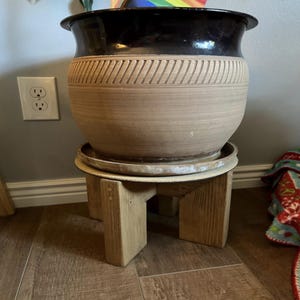 Op de afbeelding: Een grote, tweekleurige keramische plantenbak met een zwarte rand en een bruin lichaam, met een gesneden geometrisch ontwerp. De plantenbak staat op een kleine, lichtgekleurde houten plantenstandaard. De standaard heeft een eenvoudig, geometrisch ontwerp.