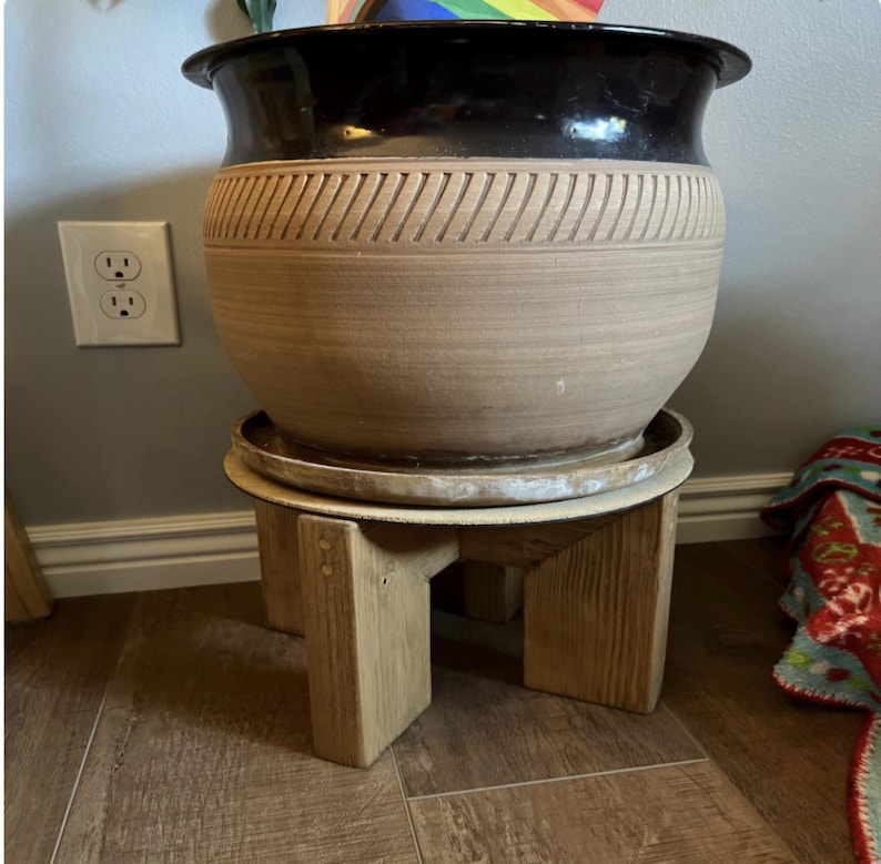 Op de afbeelding: Een grote, tweekleurige keramische plantenbak met een zwarte rand en een bruin lichaam, met een gesneden geometrisch ontwerp. De plantenbak staat op een kleine, lichtgekleurde houten plantenstandaard. De standaard heeft een eenvoudig, geometrisch ontwerp.