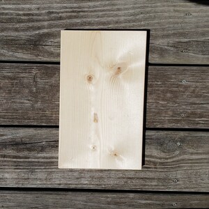 May include: A rectangular, unfinished wooden board with a light, natural wood grain. The board has visible knots and a smooth surface, suitable for crafts or DIY projects. The wood is a light beige colour.