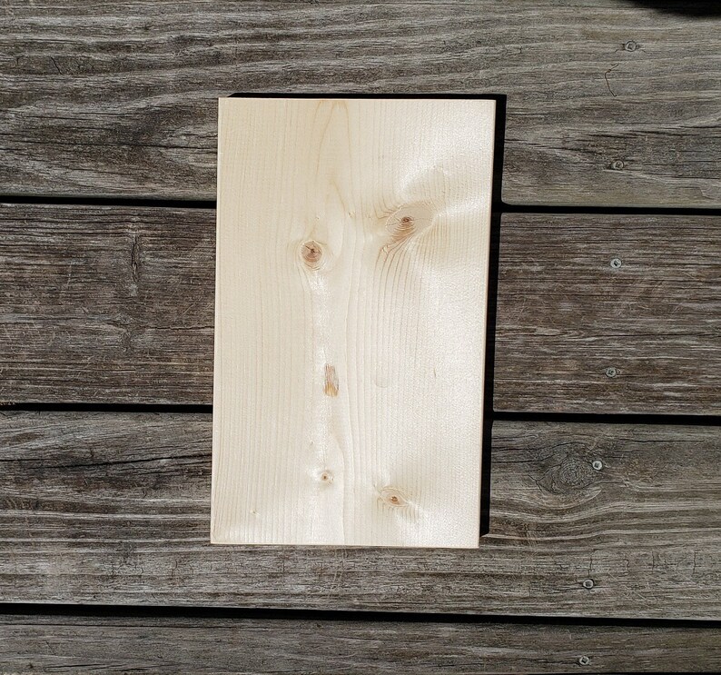May include: A rectangular, unfinished wooden board with a light, natural wood grain. The board has visible knots and a smooth surface, suitable for crafts or DIY projects. The wood is a light beige colour.