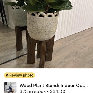 Op de afbeelding: Een grootbladige vioolbladplant in een decoratieve, gespikkelde witte pot, geplaatst op een donkerbruine houten plantenstandaard. De standaard heeft een eenvoudig, vierkant ontwerp. De plant staat binnen, bij een raam.