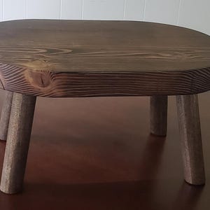 May include: A small, oval-shaped wooden stool with three legs. The stool is made of dark brown wood with a natural grain pattern.