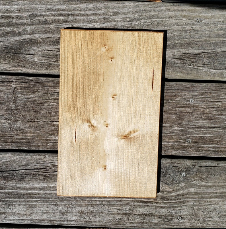 May include: A rectangular, unfinished wooden board in a light tan hue. The wood grain is visible, with several knots and small imperfections. The board is set against a weathered, grey wooden surface.