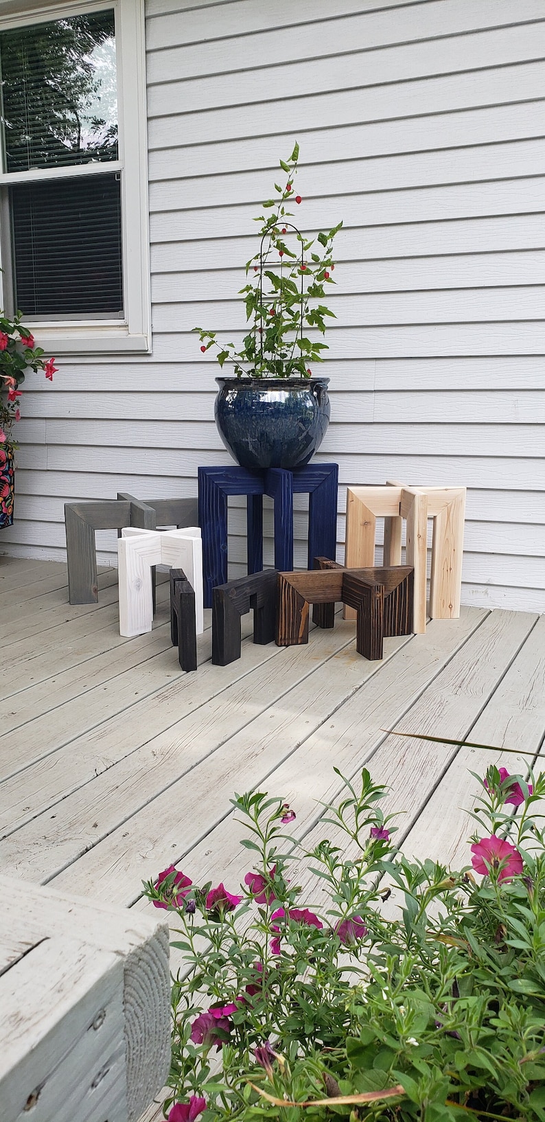 Op de afbeelding: Een verzameling houten plantenstandaards in verschillende kleuren, waaronder grijs, wit, zwart en natuurlijke houttinten. Een blauwe keramische pot met een plant staat op een standaard. De standaards worden op een houten terras getoond.