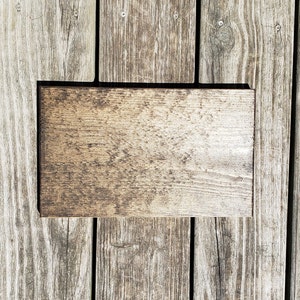 May include: A rectangular wooden sign with a dark brown border and a light brown, textured surface. The sign is placed on a weathered wooden deck, showing the natural grain and colour variations of the wood.