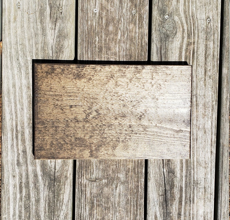 May include: A rectangular wooden sign with a dark brown border and a light brown, textured surface. The sign is placed on a weathered wooden deck, showing the natural grain and colour variations of the wood.