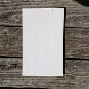 May include: A rectangular, off-white wooden board with visible wood grain and a few small knots. The board is positioned on a weathered, grey wooden surface, creating a contrast in textures and colours. The board is likely for crafts or DIY projects.