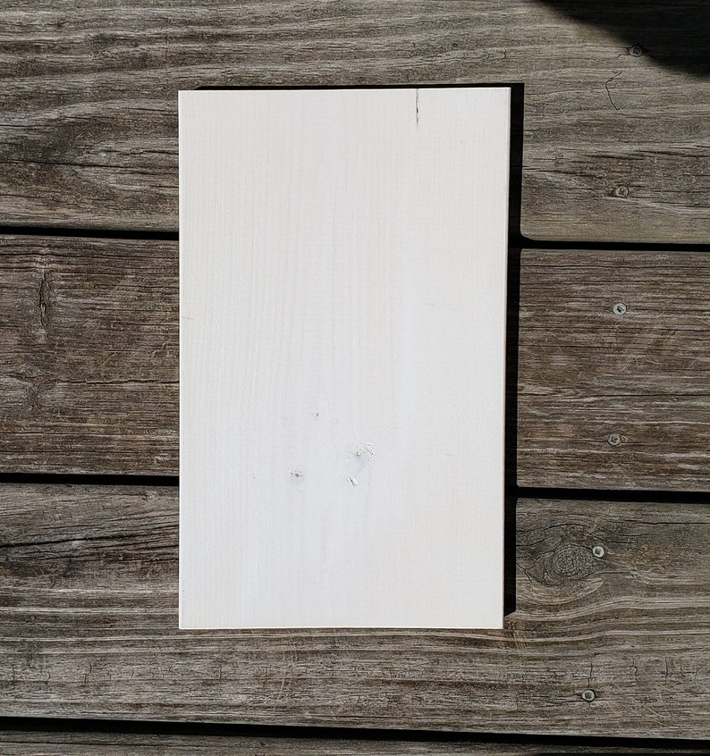 May include: A rectangular, off-white wooden board with visible wood grain and a few small knots. The board is positioned on a weathered, grey wooden surface, creating a contrast in textures and colours. The board is likely for crafts or DIY projects.