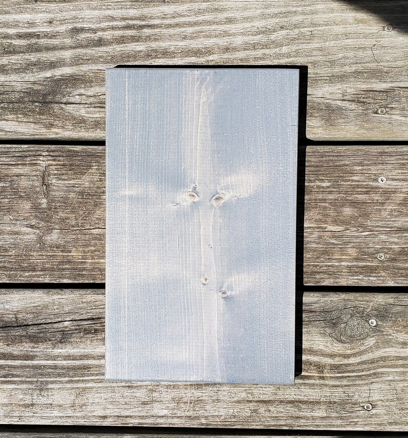 May include: A rectangular wooden sign, painted in a pale blue hue. The wood grain is visible, and the sign has a smooth surface. It is displayed on a weathered wooden surface, showing natural wood grain.