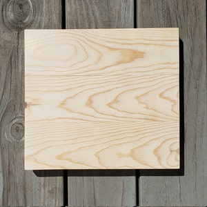 May include: A square, light-colored wooden board with visible wood grain. The board is made of three joined pieces of wood and sits on a weathered gray wooden surface.