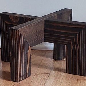Op de afbeelding: Een donkerbruine houten plantenstandaard met een X-vormige bovenkant en vier vierkante poten. De houtnerf is zichtbaar en de standaard is ontworpen om een ​​potplant te bevatten. Een decoratief item voor de inrichting.