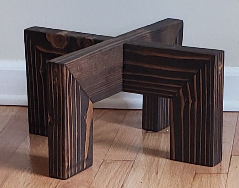 Op de afbeelding: Een donkerbruine houten plantenstandaard met een X-vormige bovenkant en vier vierkante poten. De houtnerf is zichtbaar en de standaard is ontworpen om een ​​potplant te bevatten. Een decoratief item voor de inrichting.