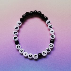 Puede incluir: Una pulsera de cuentas con las palabras "TRUE CRIME AND WINE" escritas en letras negras sobre cuentas blancas. La pulsera también presenta cuentas negras. El fondo es un degradado de rosa y morado.
