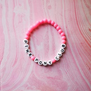 Può includere: Braccialetto di perline rosa e bianche con le parole "Pink Pony Club" scritte.