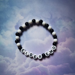 Puede incluir: Una pulsera de cuentas negras y blancas con las palabras "ELDER EMO" deletreadas en cuentas de letras blancas. El fondo es una suave mezcla de púrpura y azul nublado.
