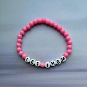 Op de afbeelding: Een roze kralenarmband met de woorden "LET THEM" gespeld in witte letterkralen. De armband is gemaakt van kleine, ronde, roze kralen en wordt gepresenteerd op een lichtblauwe en paarse achtergrond.