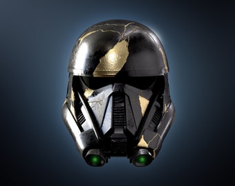 Puede incluir: Un casco negro y dorado con una superficie agrietada y desgastada. El casco tiene una visera y dos luces verdes a los lados.