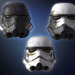 Puede incluir: Tres cascos de stormtrooper blancos y negros con detalles dorados y pintura agrietada. Los cascos están dispuestos en formación triangular.