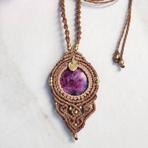 May include: A macrame necklace featuring a round, deep magenta and purple stone pendant encased in brown cord. The design includes intricate knotted patterns, small gold-toned metallic beads, and a small gold-toned leaf-shaped charm at the top.